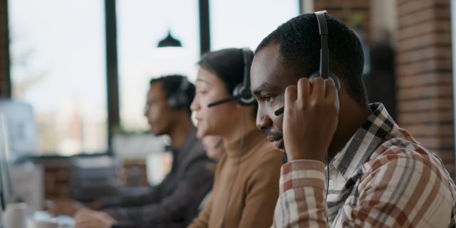 Male worker answering telework phone at call center, using audio headset and microphone. Young man working at customer care in disability friendly office. Handheld shot. Close up.